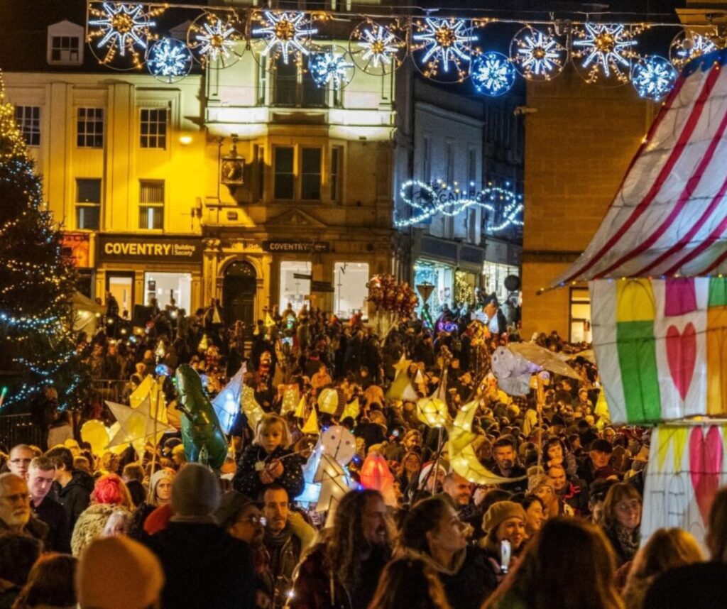 A busy street, Frome Market Place at Christmas. Gold, white and blue Christmas lights above the street and on the Christmas tree. A crowd carrying willow and tissue lanterns lit from inside