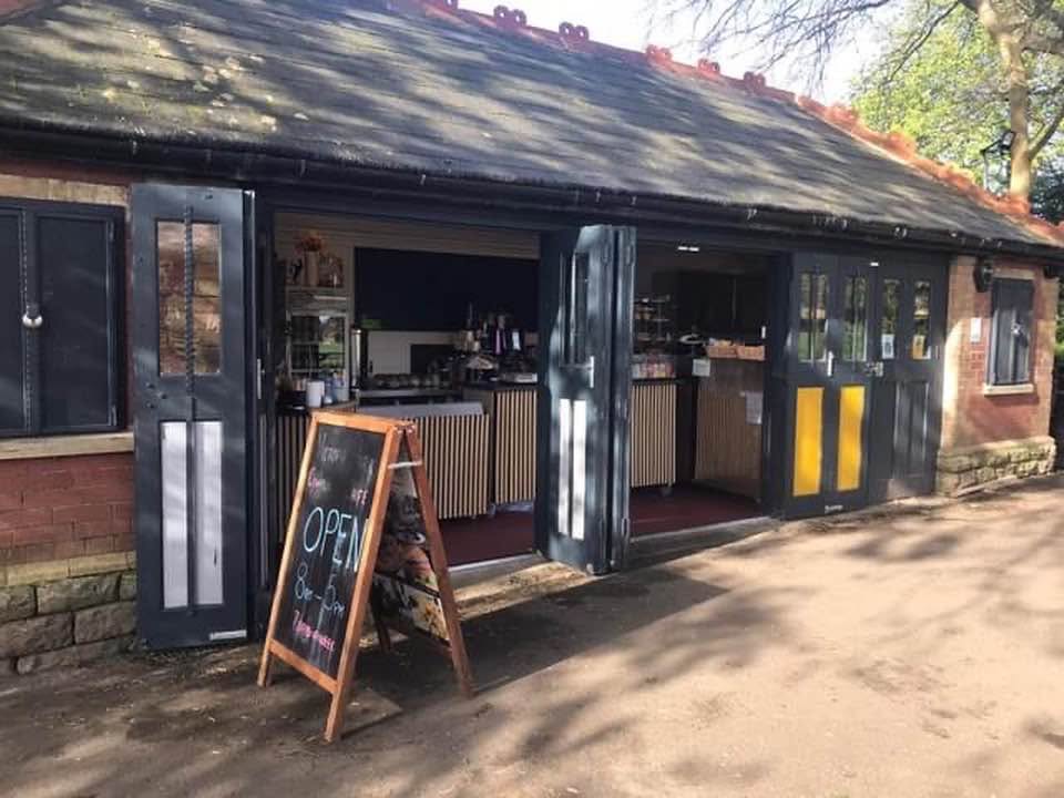 The community cafe in Victoria Park, a red brick building with wooden green and yellow shutters running along the front. The shutters are open, an A-board says 'open' and the sun is shining.
