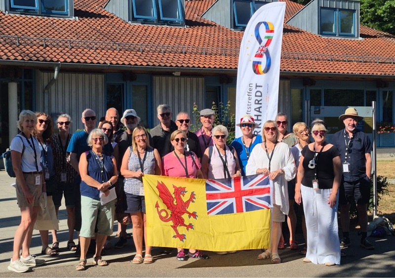 A large group smiling in the sunshine with a Somerset/Union Jack Flag held in front and a feather banner for Frome's twon town of Murrhardt, germany in the background.