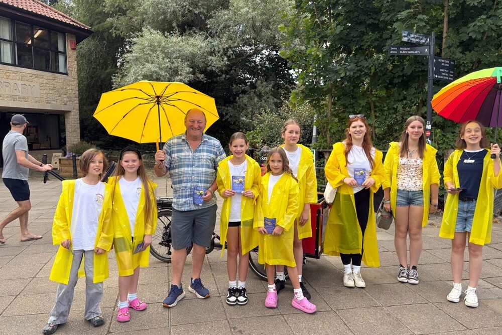 Mayor Mark Dorrington with young cast members from a production of Singin in the Rain, wearing yelow rain macs and carrying brightly coloured umbrellas.