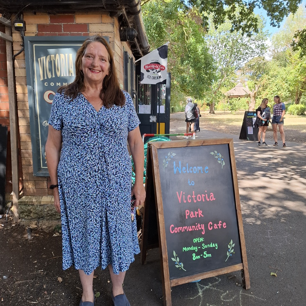 Cafe manager Caroline Blake, smiling next to a 'Welcome to Frome Community Cafe' sign, with the park and cafe in the background.